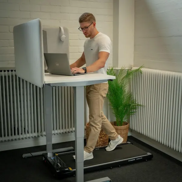 FitNord SlimWalk One -kävelymatto korkeussäädettävän työpöydän alla. Kokoontaitettava walking pad sopii kotitoimistoon ja päivittäiseen kävelyyn.